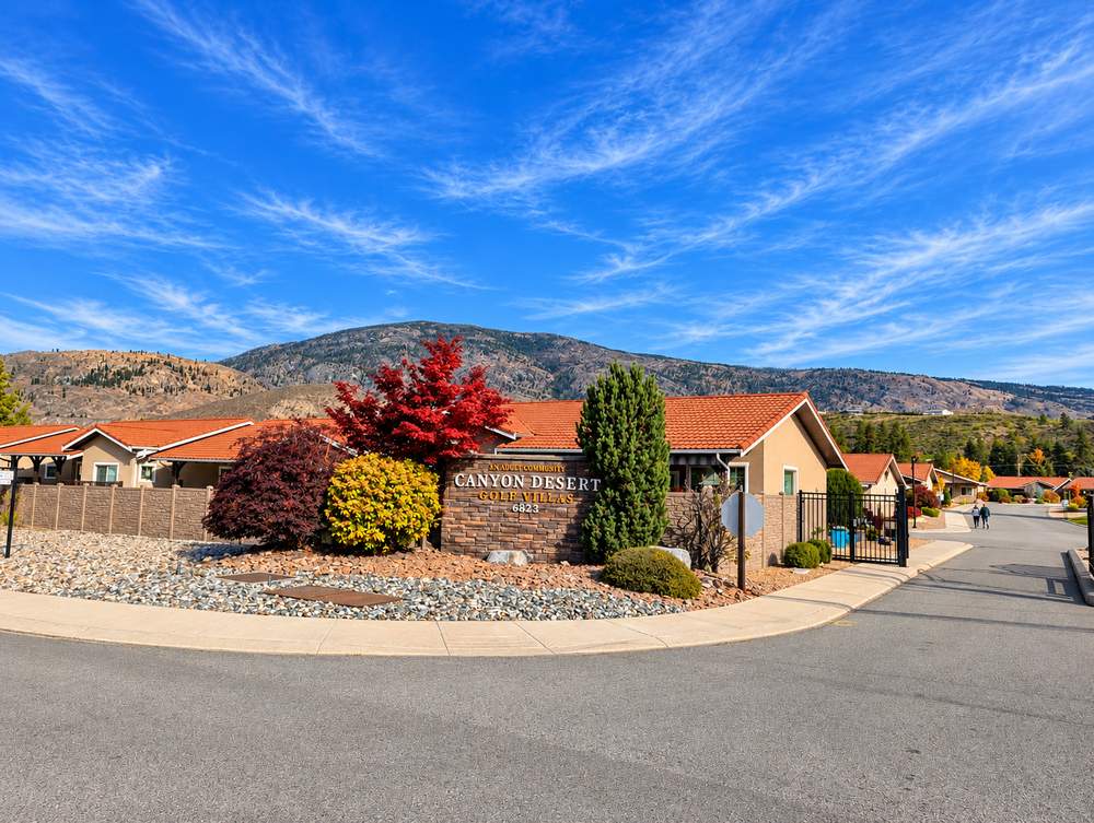 Canyon Desert Golf Villas stone-faced entrance sign reading An Adult Community, Canyon Desert Golf Villas, 6823, with tile-roofed homes, a black gate and hills behind