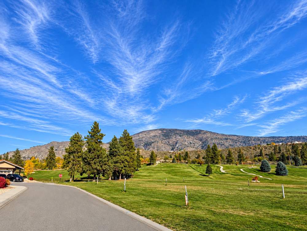 Golf course fairway with tall pines and a bare hillside behind, seen from a road at Canyon Desert Golf Villas